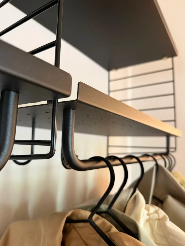 String Regal als Garderobe von unten fotografiert, mit schwarzer Metallstange, eingehängten Kleiderbügeln und Holzboden auf schwarzen String Leitern