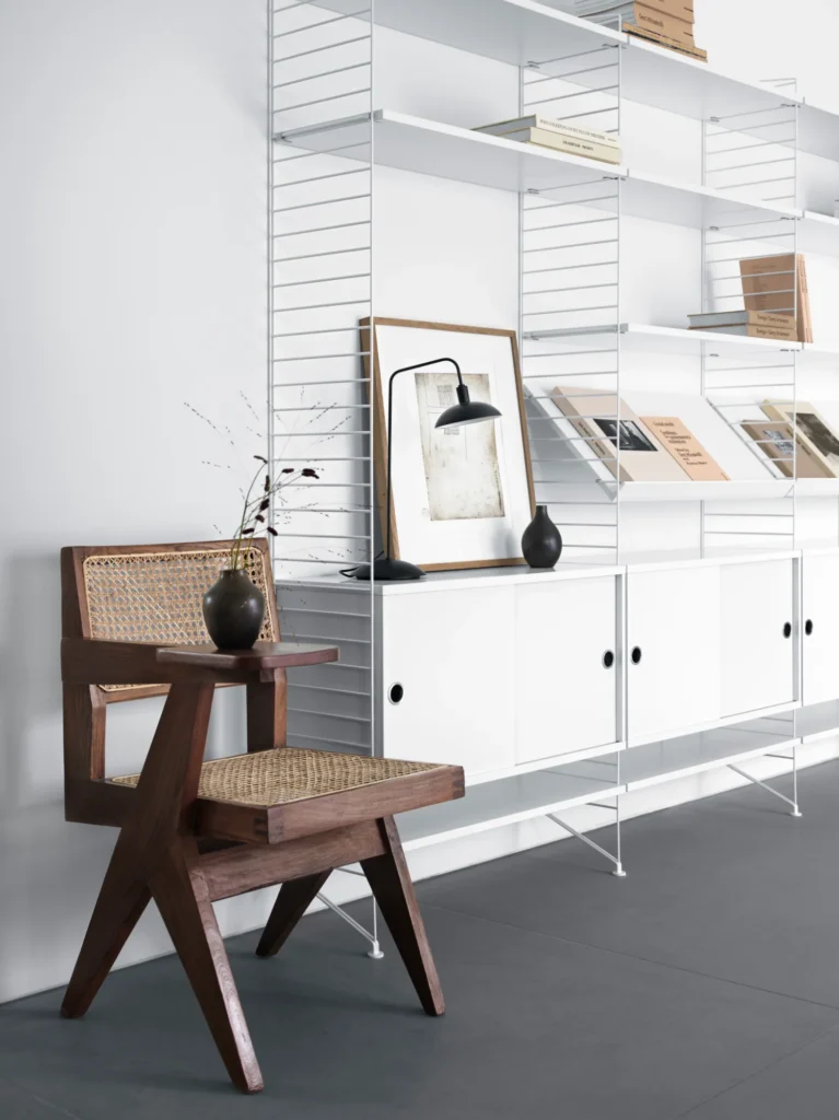 Weißes String System mit Schiebetürschränken und Zeitschriftenböden, davor ein Holzstuhl mit Geflecht